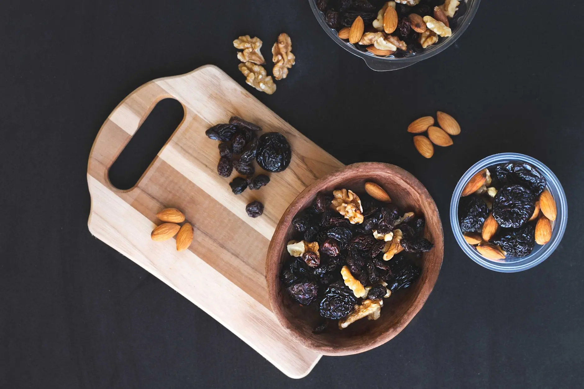 A top-down view of mixed dried fruits and nuts in bowls on a black surface with a wooden chopping board