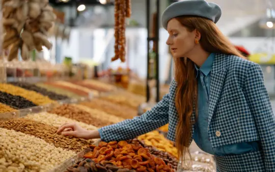 Woman in stylish outfit selecting dried fruits and nuts at a market display.
