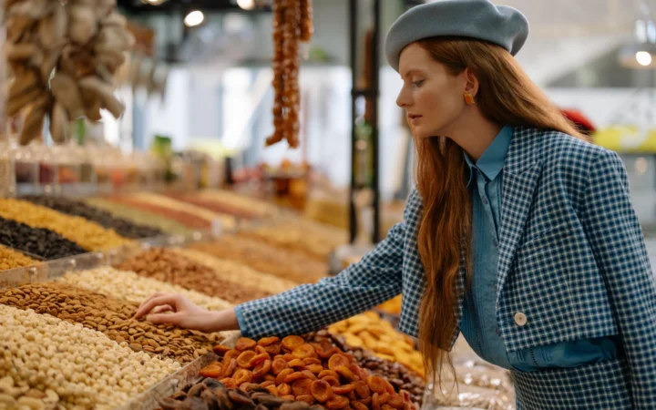 Woman in stylish outfit selecting dried fruits and nuts at a market display.