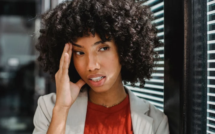 Businesswoman with curly hair expresses stress by her office window, touching her forehead.