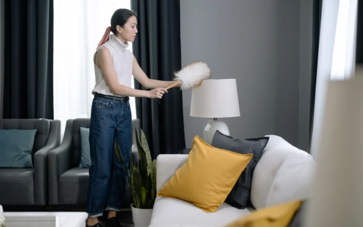 Asian woman dusting a lampshade in a modern living room with yellow pillows.
