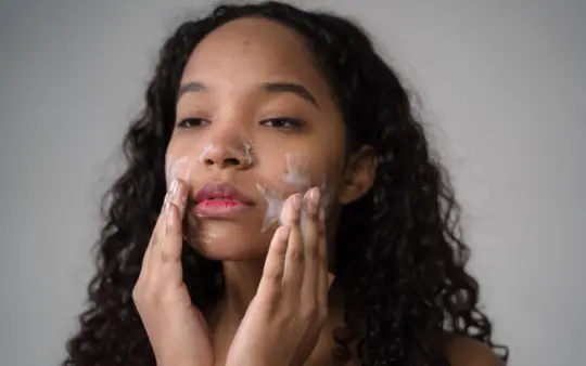 African American female with long dark curly hair washing cheeks with facial foam on gray background
