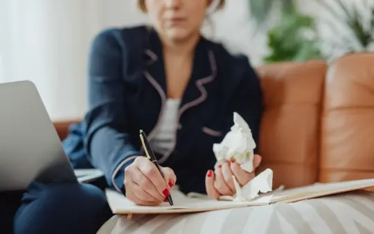 Woman writing in a notebook with tissues nearby, suggesting remote work or illness at home.