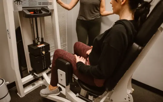 Woman performs exercises on a hip abductor machine with guidance from a trainer.