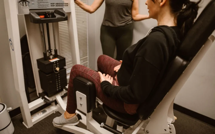 Woman performs exercises on a hip abductor machine with guidance from a trainer.