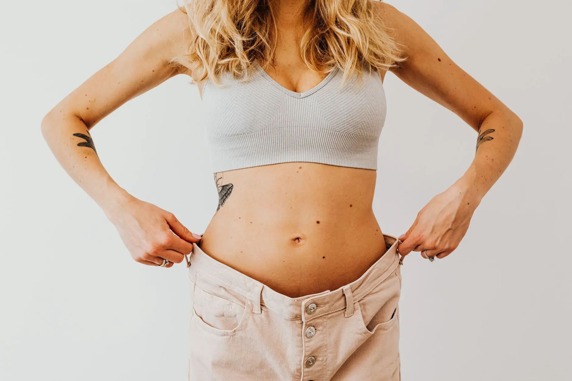 A woman displaying weight loss by wearing oversized pants and crop top, showing tattoos