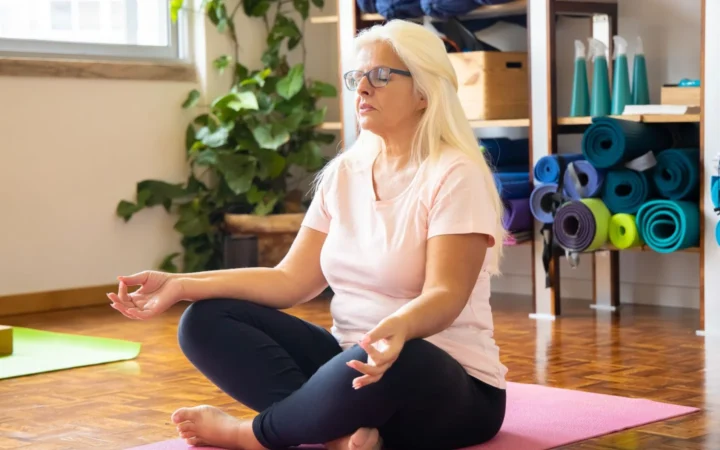 Adult woman sits cross-legged on a mat meditating in a serene yoga studio environment.