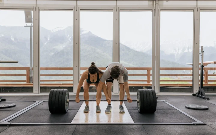 Couple lifting a heavy barbell together in a gym with mountain views.