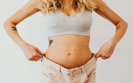 Woman displaying weight loss by wearing oversized pants and crop top, showing tattoos.