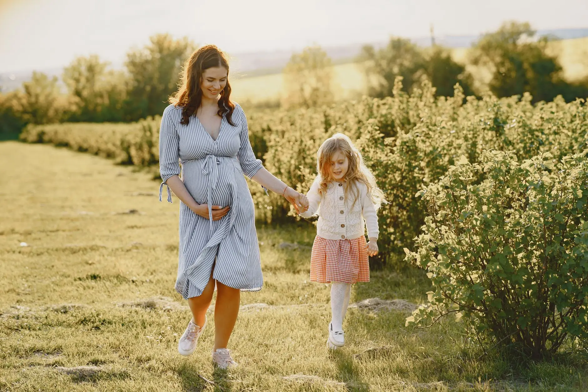 A pregnant mother holds her daughter's hand while walking in a sunny field surrounded by greenery