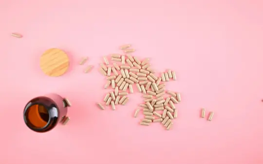 Brown bottle with scattered herbal supplement capsules on a pastel pink background.