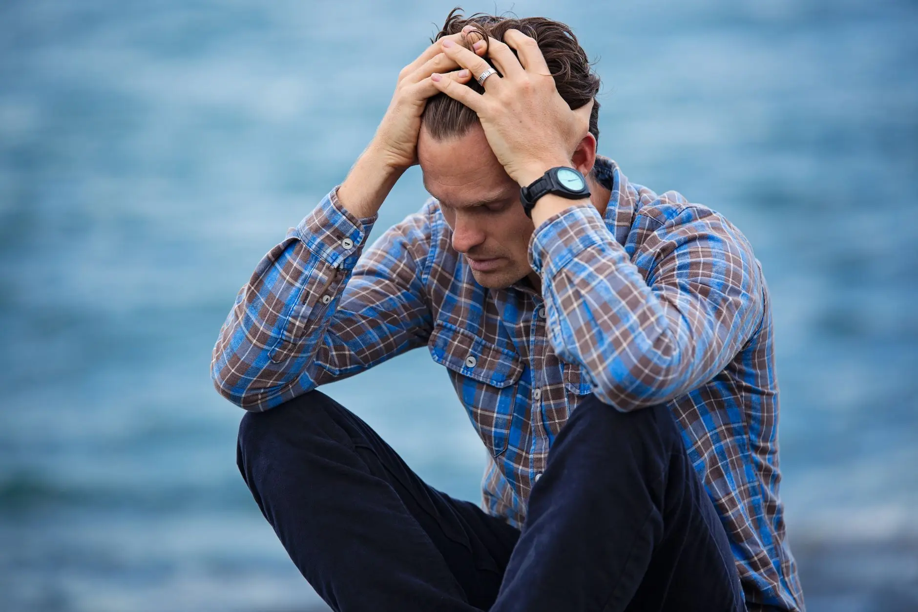 A man in a plaid shirt sits by the water looking distressed, symbolizing stress