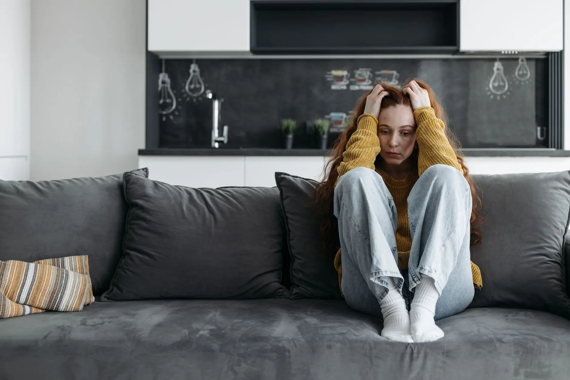 A woman in a yellow sweater sits on a couch, looking sad and pensive, indoors
