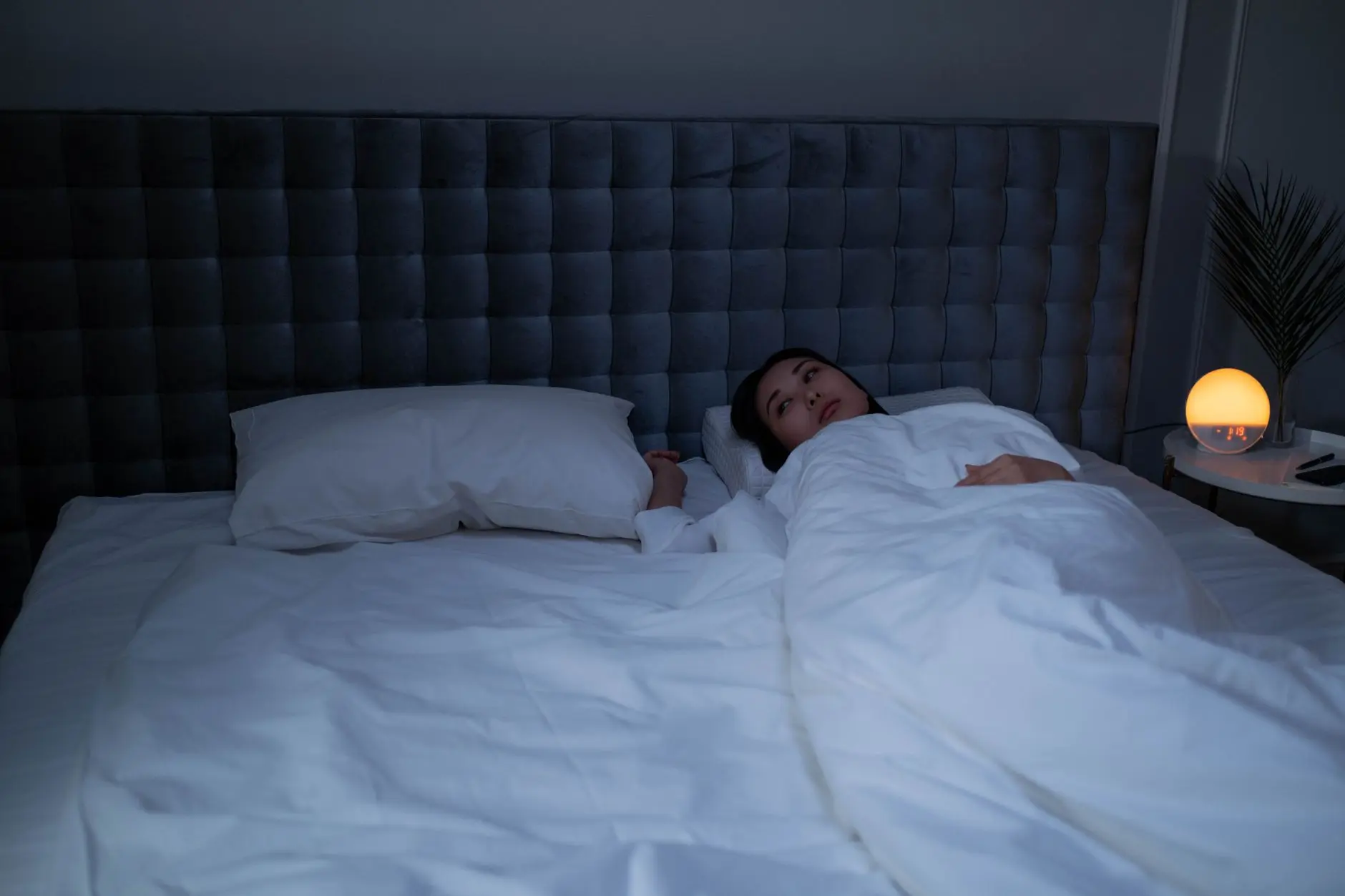 An Asian woman lying awake under soft white bedding in a dimly lit modern bedroom