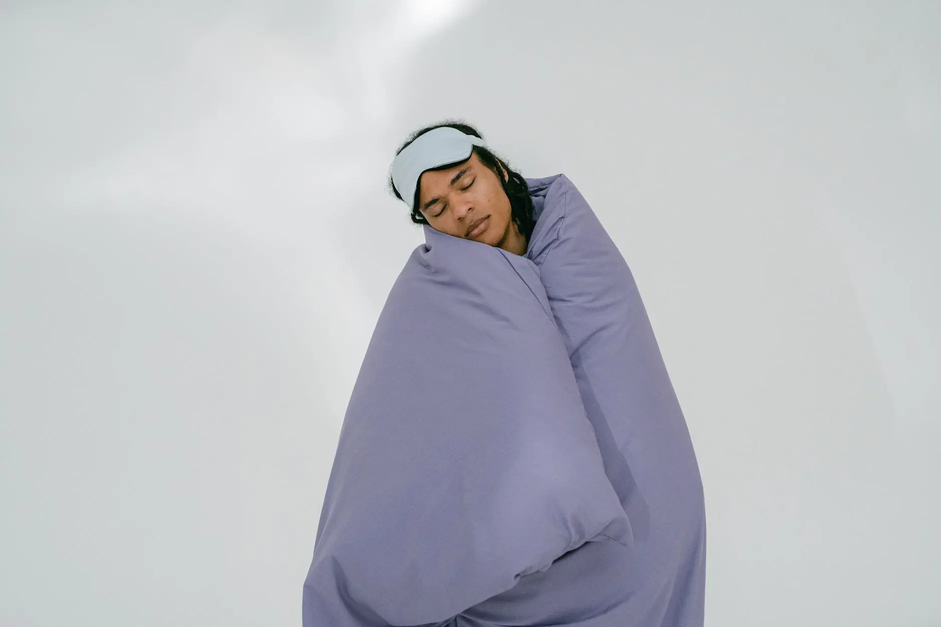 A person peacefully sleeping with an eye mask, wrapped in a purple blanket on a white background