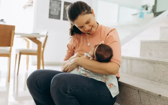 Woman smiling and cradling baby on stairs indoors, portraying warmth and family bonding.