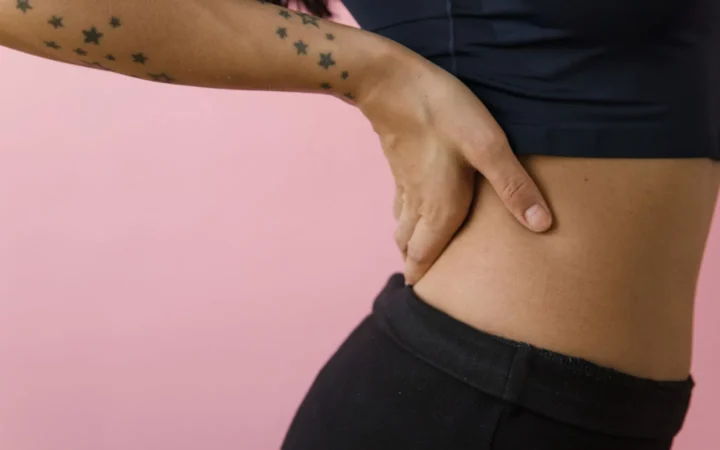 Side view of a tattooed hand on hip against a pink background in a studio setting.