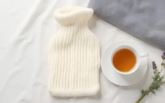 Seen from directly above, filling the frame, on a pale linen cloth: a hot water bottle in a cream knitted cover, a cup o, generated illustration