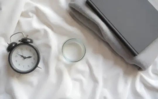 Seen from directly above, filling the frame, on a pale linen sheet: an analogue alarm clock with a plain face, a closed , generated illustration