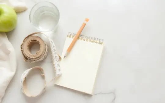 Seen from directly above on a pale stone worktop, a coiled cloth tape measure, a glass of water, a small open notebook w, generated illustration