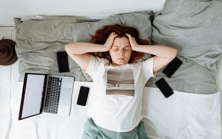 Distressed woman lying in bed surrounded by digital devices, showing signs of stress.