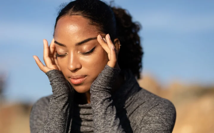 Close-up of a woman in a gray sweater holding her temples, experiencing a headache outdoors in the sunlight.