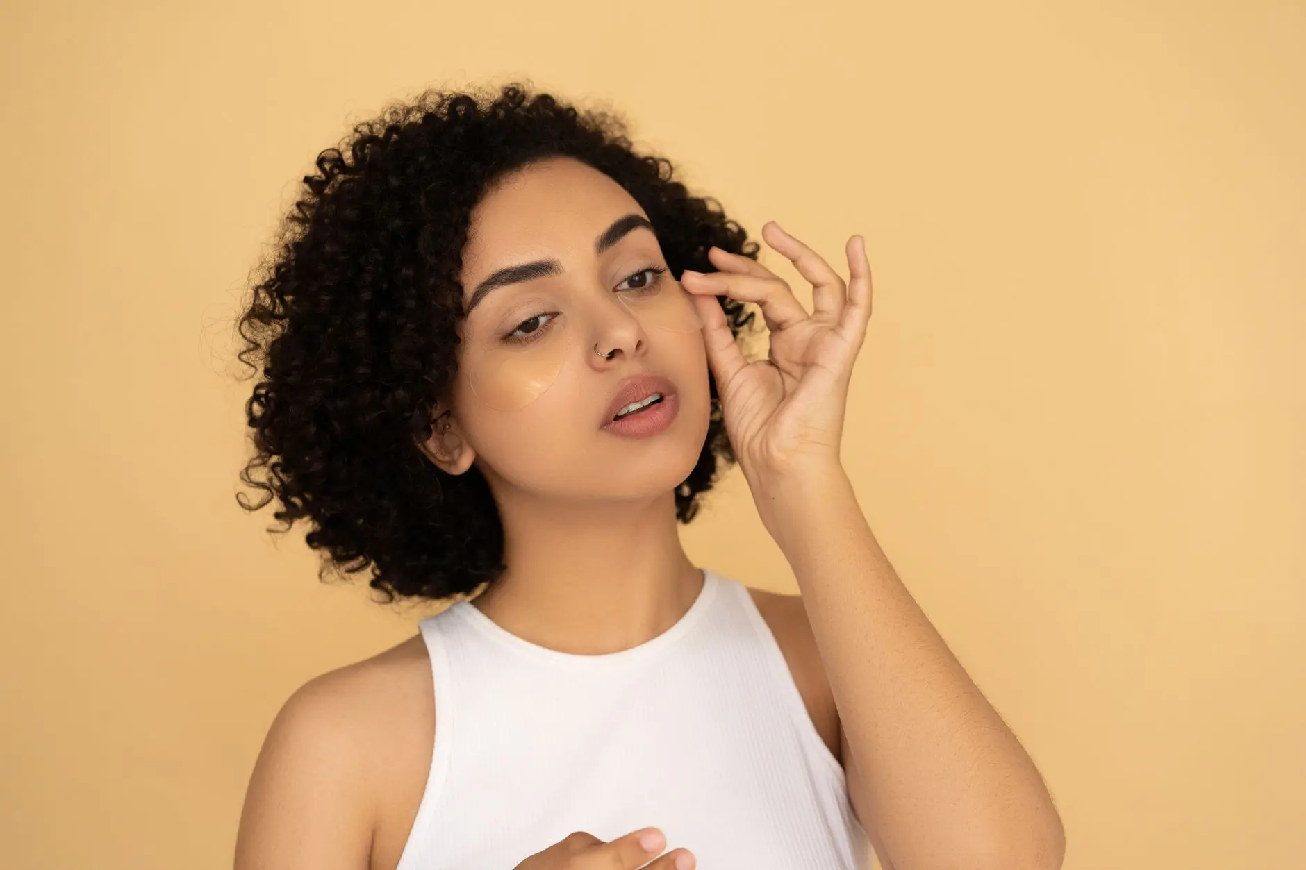 A woman with curly hair applies under eye masks against a neutral background