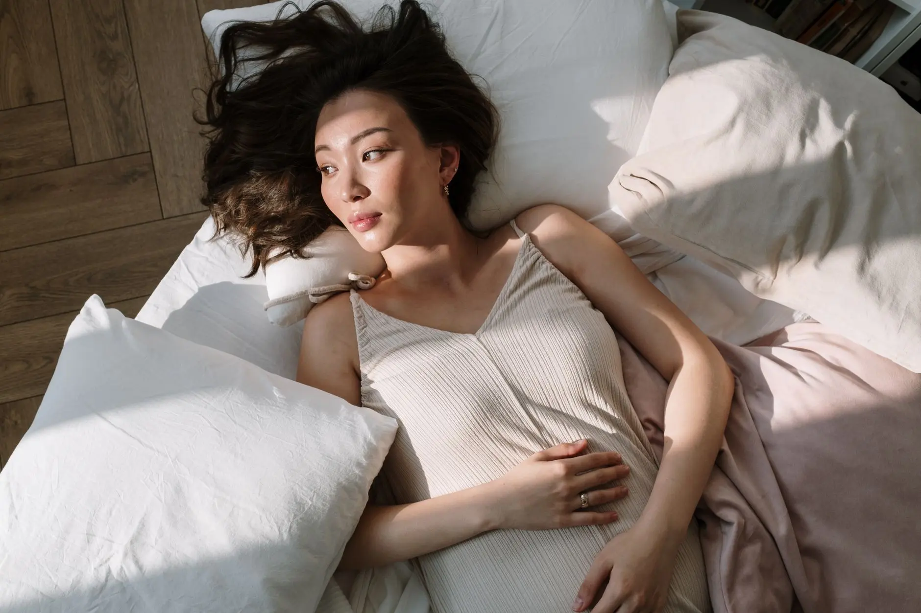 Asian woman in sleepwear lying on bed with hand on belly, enjoying morning sunlight