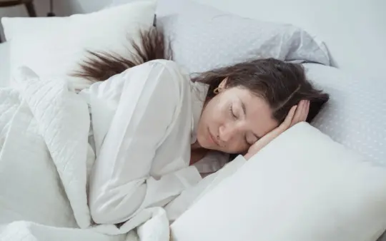 Serene woman rests comfortably in bed with soft pillows and blankets, embracing relaxation.