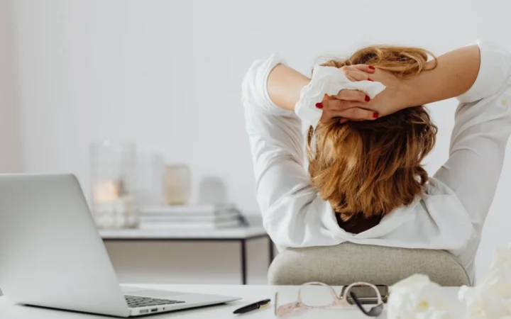 Adult woman in white takes a break at her desk, holding a tissue, with a laptop and eyeglasses nearby.