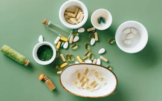 Top view of various herbal and pharmaceutical supplements in ceramic bowls on a green background.
