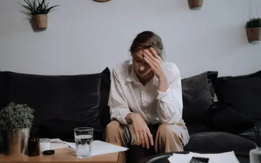 Woman experiencing stress and headache while sitting on a sofa at home, surrounded by papers.