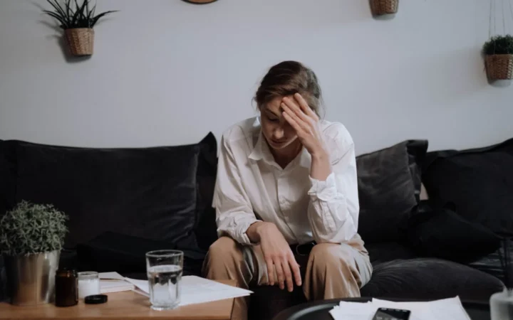 Woman experiencing stress and headache while sitting on a sofa at home, surrounded by papers.