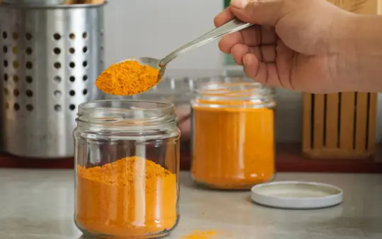 Close-up of turmeric powder being scooped into a jar in a kitchen with utensils.