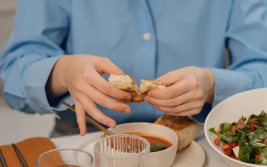 Woman tearing bread over a bowl of soup, wearing a blue shirt, indoors.