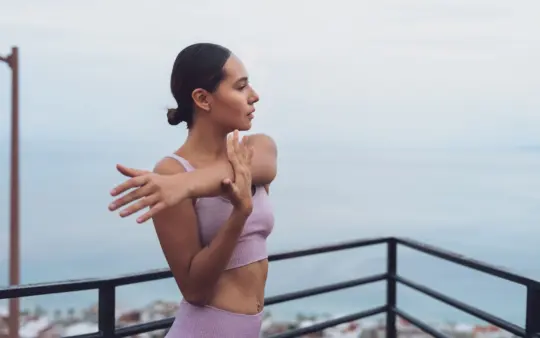 Young woman in workout attire stretching on a balcony with a sea view.