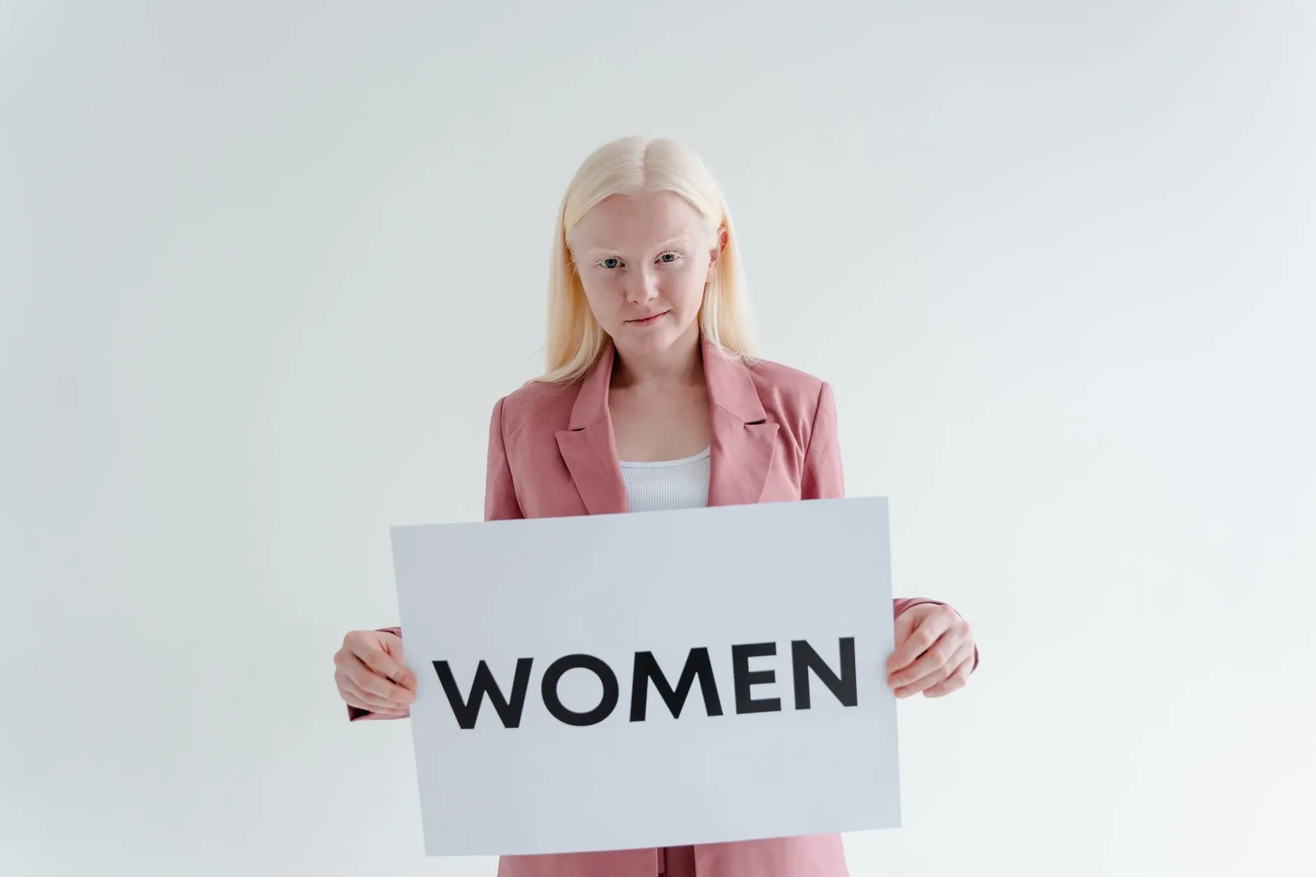 Confident woman in pink blazer holds a sign reading 'Women' on a white background