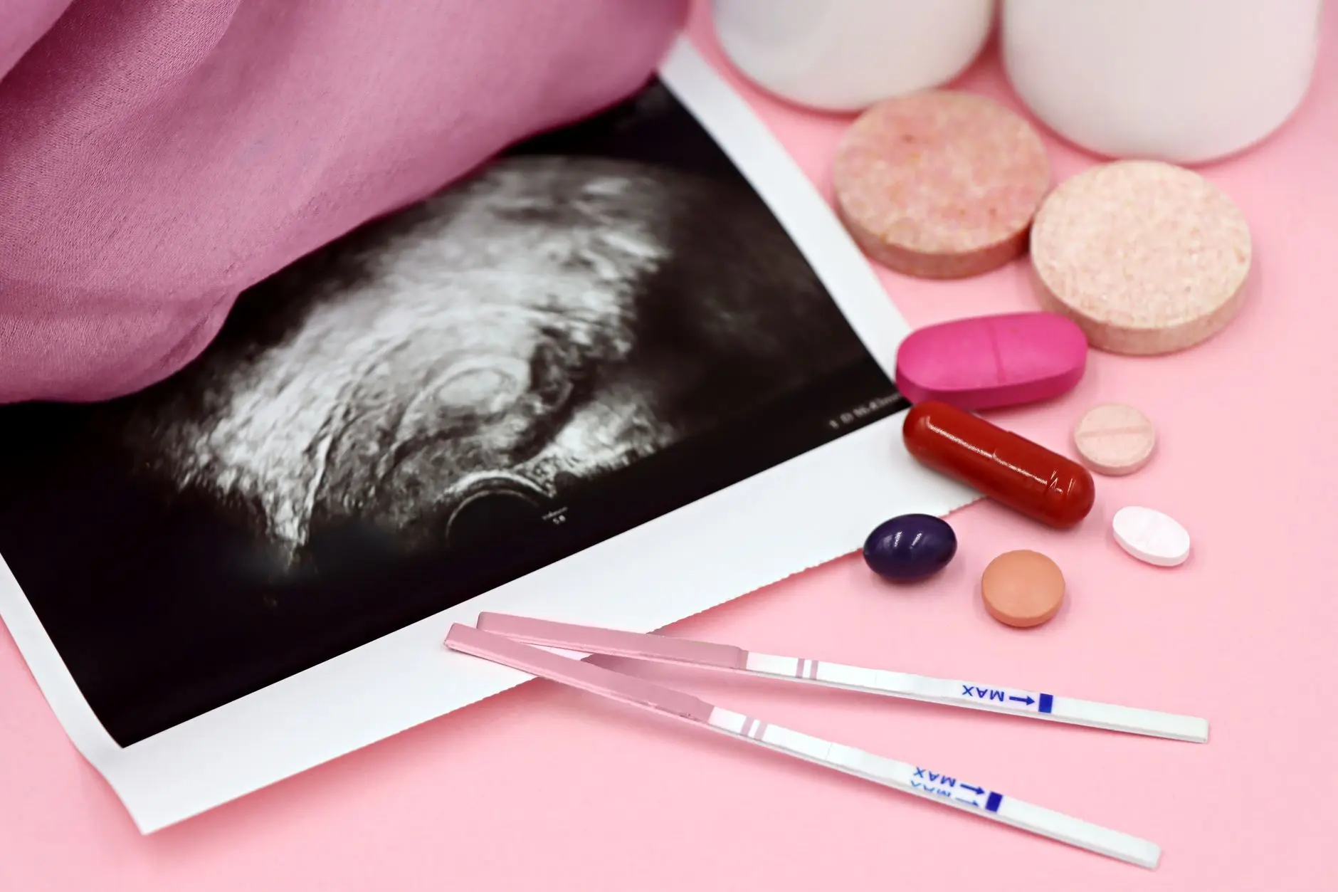 A closeup of pregnancy tests and an ultrasound with supplements on a pink background