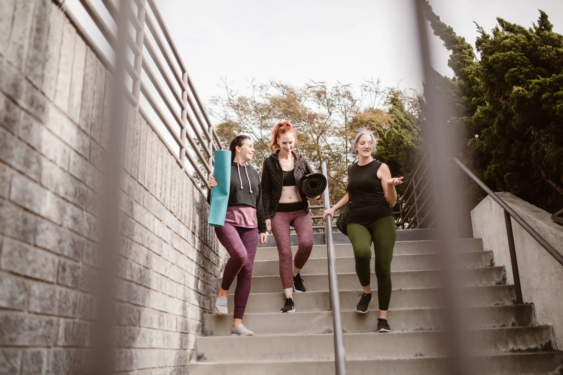 Three women walking down steps outdoors, carrying yoga mats, enjoying a fitness conversation