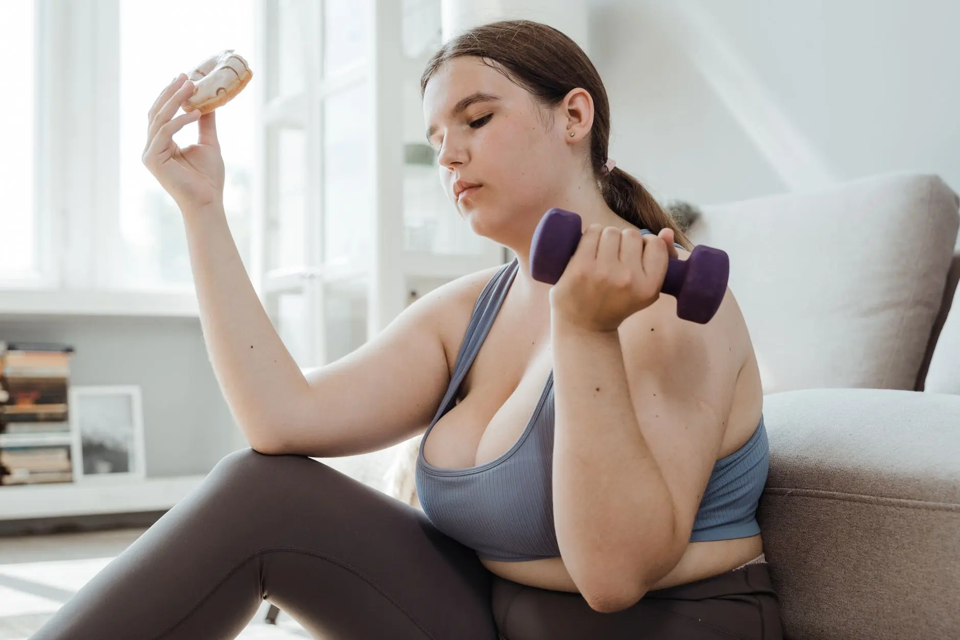 A plus size woman in activewear balancing a dumbbell and a donut, symbolizing fitness and indulgence