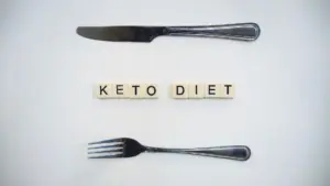 Ketogenic diet weight loss plateau: top view of a knife, fork, and scrabble tiles spelling 'Keto Diet' on a white surface