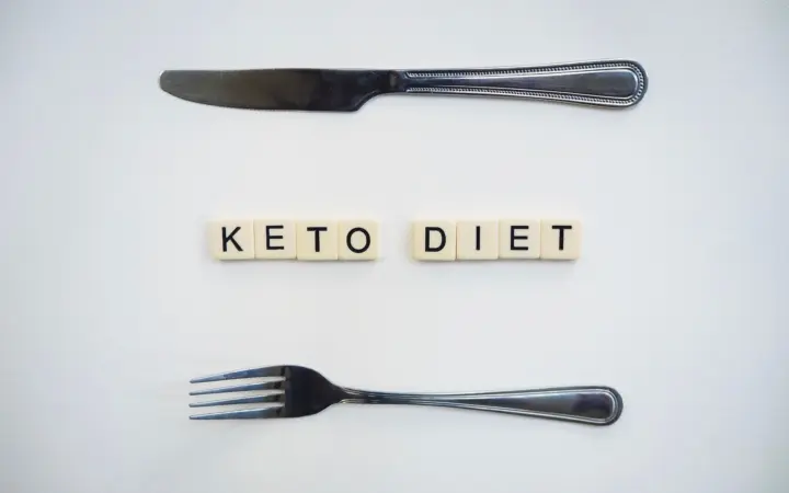 Ketogenic diet weight loss plateau: top view of a knife, fork, and scrabble tiles spelling 'Keto Diet' on a white surface