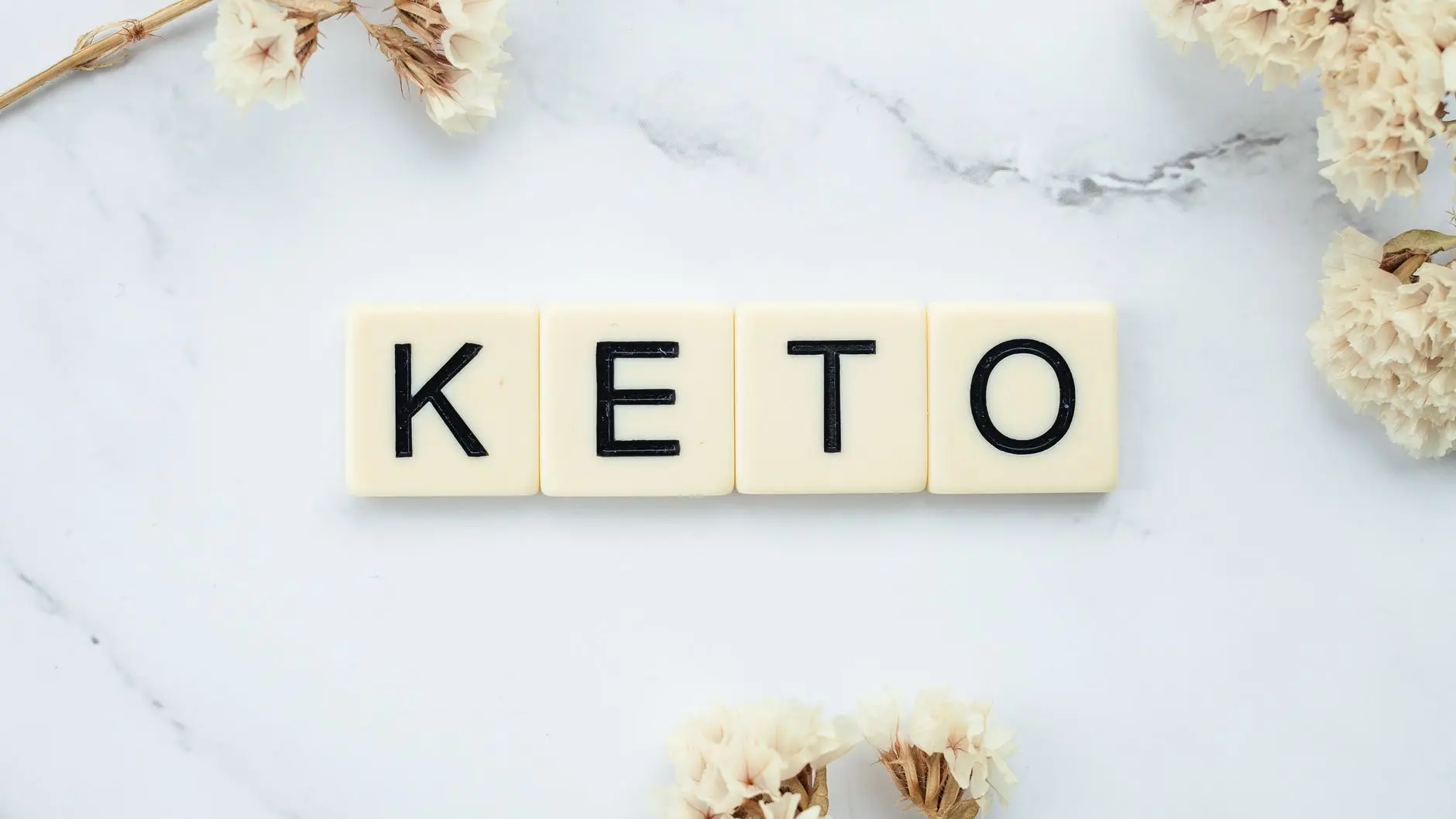 Scrabble tiles spelling 'Keto' on a marble surface with white flowers, symbolizing a ketogenic lifestyle