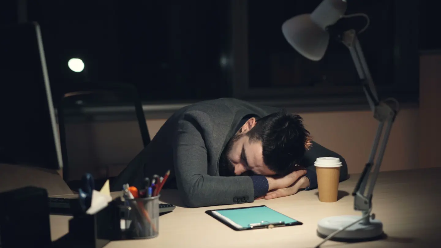 Sleep hygiene for night shift workers: man in office suit sleeping on desk with coffee and tablet nearby, symbolizing work