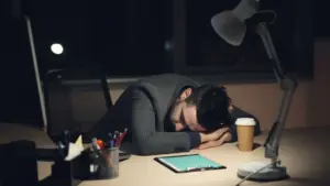Sleep hygiene for night shift workers: man in office suit sleeping on desk with coffee and tablet nearby, symbolizing work