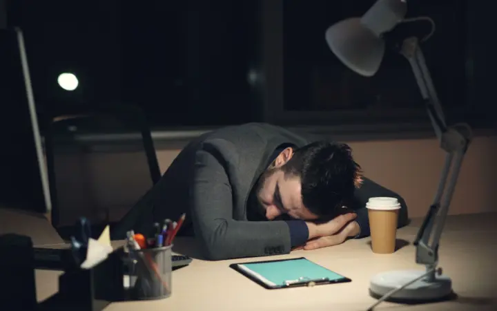Sleep hygiene for night shift workers: man in office suit sleeping on desk with coffee and tablet nearby, symbolizing work