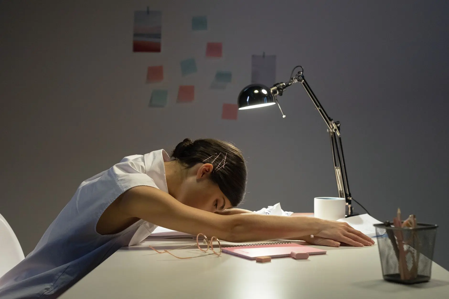 A tired woman rests her head on an office desk under a lamp, symbolizing workplace fatigue