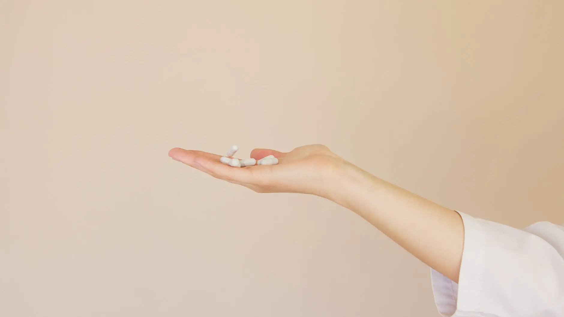 Side view of crop unrecognizable female pharmaceutist in white uniform with pile of white painkillers on palm against l