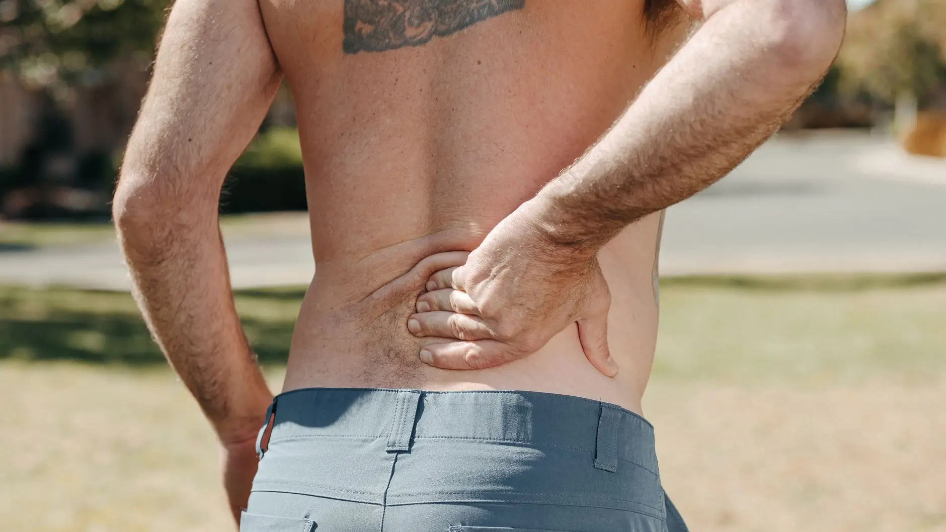 Shirtless man holding his lower back outdoors indicating pain or discomfort